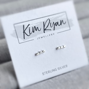 Puede incluir: Pendientes de plata de ley, cada uno con tres pequeñas cuentas circulares conectadas. Se exhiben en una tarjeta blanca con el texto "Kim Ryan Jewellery" y "STERLING SILVER". El fondo es un gris suave y borroso.