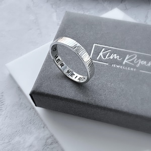 May include: A silver ring with the words "HER WEIRD" engraved inside. The ring has a textured surface and is displayed on a grey jewellery box with the text "Kim Ryan Jewellery".