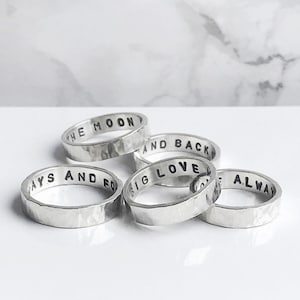 Può includere: Sono presentati cinque anelli d'argento con texture martellate. Ogni anello ha una frase incisa diversa: "TO THE MOON", "AND BACK", "ALWAYS AND FO", "BIG LOVE" e "ALWAYS". Gli anelli sono su una superficie bianca.