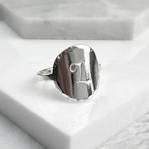May include: A silver ring with a hammered, oval-shaped top featuring the letter "L" in a cursive font.