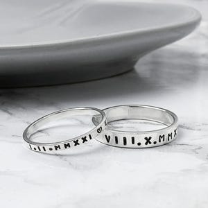May include: Two silver rings with engraved Roman numerals and dates. One ring has a heart symbol. The rings are on a marble surface with a blurred white plate in the background. The rings are likely wedding or anniversary bands.