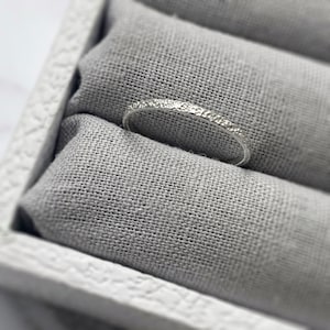 May include: A silver ring with a textured surface, displayed on a grey fabric cushion within a white box. The ring has a subtle, hammered texture, adding detail to the simple band. The ring is a delicate piece of jewellery.