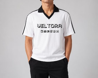 Può includere: Una polo bianca a maniche corte con colletto a V nero e strisce nere lungo le spalle. Sul petto è presente la scritta VELTORA MILANO in un font nero pixelato. La maglia è abbinata a pantaloni neri a tinta unita.