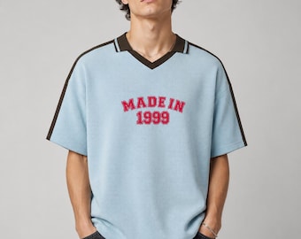 Puede incluir: Un polo de manga corta azul claro con cuello en V marrón oscuro y rayas marrones oscuras a juego en los hombros. El pecho presenta el texto bordado en rojo "MADE IN 1999" con una tipografía llamativa de estilo universitario.