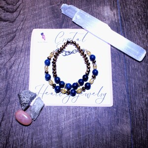 May include: A handmade bracelet with dark blue and gold beads, and a second bracelet with brown and gold beads, rests on a white card with the words "Crystal Healing Jewelry". A white selenite wand and several crystals are also present.