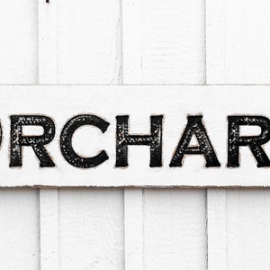 May include: White wooden sign with black lettering that reads "ORCHARD".