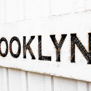 May include: White wooden sign with the word "BROOKLYN" in black lettering. The letters are raised and have a textured surface.
