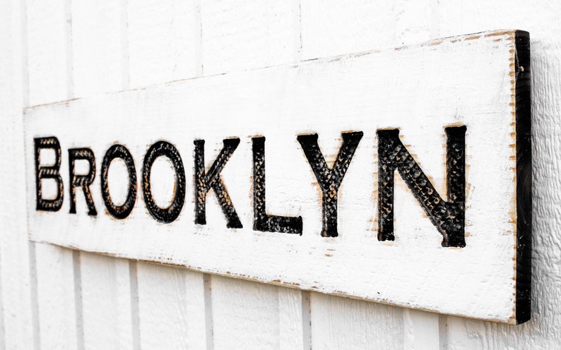 May include: White wooden sign with the word "BROOKLYN" in black lettering. The letters are raised and have a textured surface.