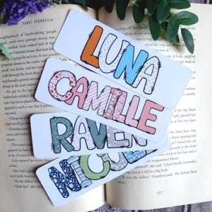 May include: Four personalized name bookmarks are displayed, each with a different name: Luna, Camille, Raven, and Nico. The names are in large, colorful, bubble letters with various patterns. The bookmarks are on top of an open book.