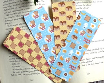 May include: Set of four colorful bookmarks featuring different breakfast food patterns. The bookmarks are printed on a light blue, yellow, and light blue background. The patterns include pancakes, bacon, eggs, toast, and orange juice.