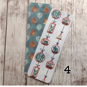 May include: Two bookmarks, one with a teal background and a pattern of copper-colored leaves, and the other with a white background and a pattern of copper-colored hanging terrariums with plants.
