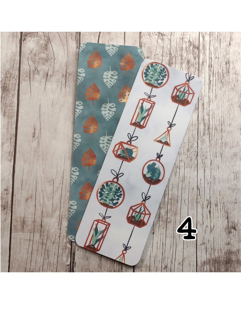 May include: Two bookmarks, one with a teal background and a pattern of copper-colored leaves, and the other with a white background and a pattern of copper-colored hanging terrariums with plants.