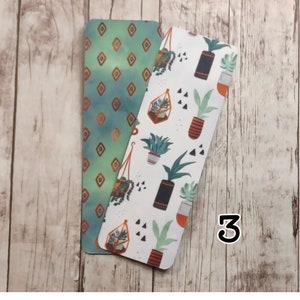 May include: Two bookmarks, one with a green and gold geometric pattern and the other with a white background and a pattern of potted plants, succulents, and geometric shapes. The number "3" is written on the wooden surface.
