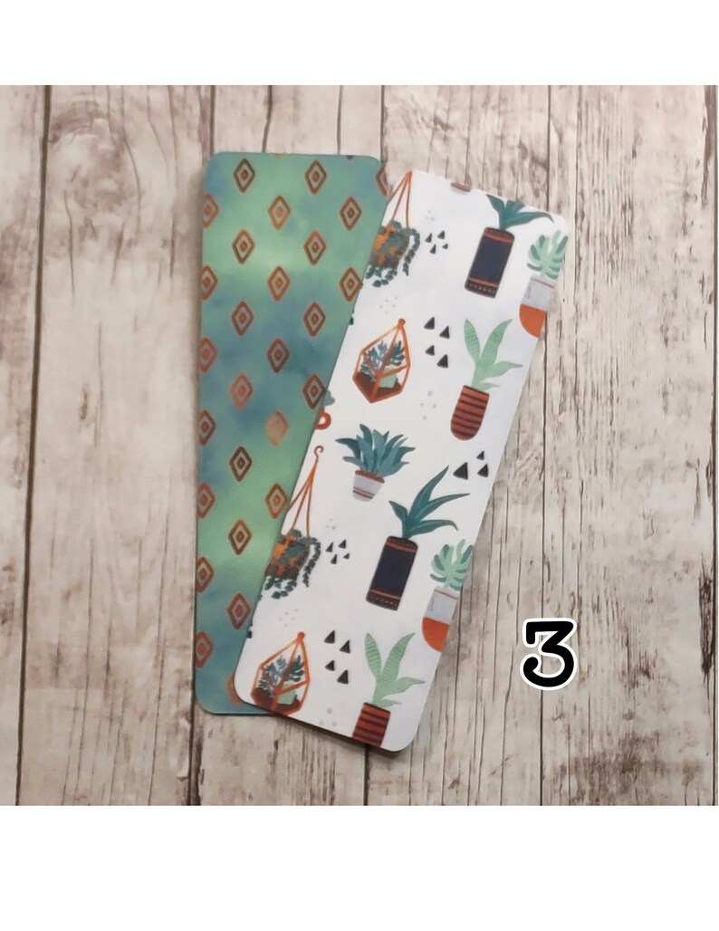 May include: Two bookmarks, one with a green and gold geometric pattern and the other with a white background and a pattern of potted plants, succulents, and geometric shapes. The number "3" is written on the wooden surface.