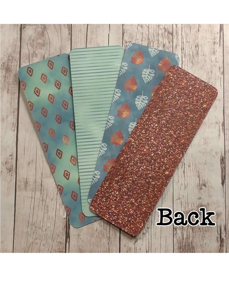 May include: Four different bookmark designs. One bookmark has a geometric pattern with copper accents on a blue background. Another bookmark has a green and white striped pattern. A third bookmark has a blue background with white leaves. The fourth bookmark has a copper glitter background. The back of the bookmarks is visible.
