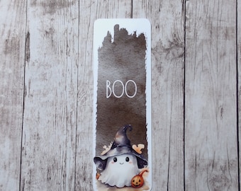 May include: A white bookmark with a watercolor illustration of a ghost wearing a witch hat and holding a small pumpkin. The background is a dark gray watercolor wash. The text "BOO" is written in white on the bookmark.