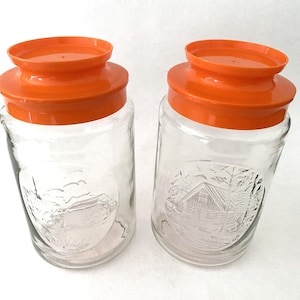 May include: Two clear glass jars with orange lids. The jars have an embossed design of a cabin in the woods.