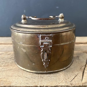 May include: A round, antique brass box with a hinged lid and a clasp closure. The box has a handle on top and sits on a wooden surface. The brass has a warm, aged patina.