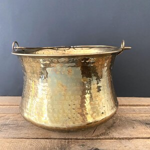 May include: A hammered brass cauldron with two small handles. The antique pot has a wide, slightly flared rim and a rounded body. The surface is textured with a dimpled pattern, reflecting light. The background is a dark blue wall and wooden planks.