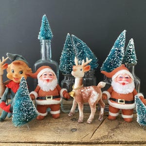 May include: A collection of vintage Christmas decorations, including Santa Claus figures, reindeer, and bottle brush trees. The Santa figures wear orange suits with black belts. The reindeer have gold bells. The trees are teal.