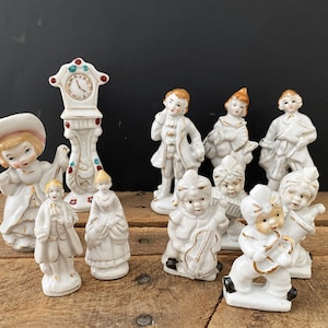 May include: A collection of ten white porcelain figurines, including a clock tower, a girl in a dress, a boy in a suit, and several musicians playing instruments. The figurines are decorated with gold and red accents.
