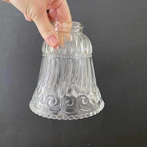 May include: Clear glass lampshade with a bell shape, featuring vertical ribbed detailing and a decorative swirl pattern at the bottom. The lampshade is held by a hand against a dark background.