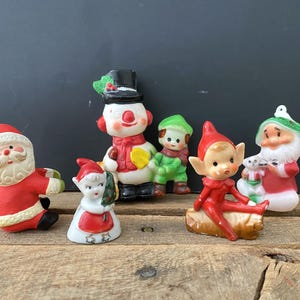 May include: A collection of vintage Christmas figurines, including Santa Claus, a snowman, elves, and a small child. The figurines are painted in bright colors and are arranged on a wooden surface.