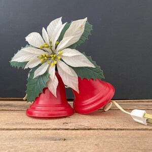 May include: Two bright red plastic bells decorated with a large white poinsettia flower featuring yellow centers and green leaves. The bells are connected by a white electrical cord with a standard two-prong plug extending from the side.