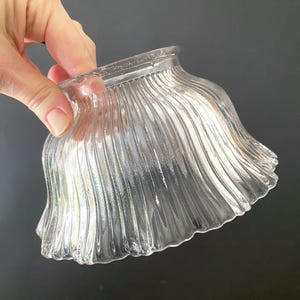 May include: A clear glass lampshade with a wavy, ribbed design. The shade has a flared, ruffled edge and a smooth, rounded top. The glass is transparent, allowing light to pass through.