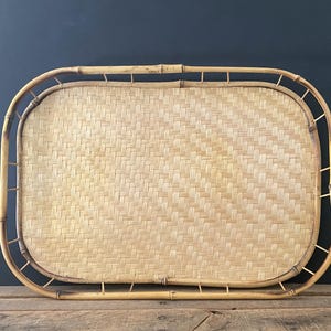 May include: Rectangular bamboo serving tray with a woven center. The tray has a natural light brown color and a curved bamboo frame. The woven center is a lighter shade of brown. The tray is sitting on a wooden surface.