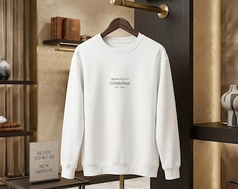 Op de afbeelding: Een wit sweatshirt met lange mouwen en een ronde hals, voorzien van de zwarte tekst "PROMOTED TO Grandma EST. 2027" in het midden op de borst. Het kledingstuk heeft een geribbelde halslijn, boorden en zoom en hangt aan een houten kledinghanger.