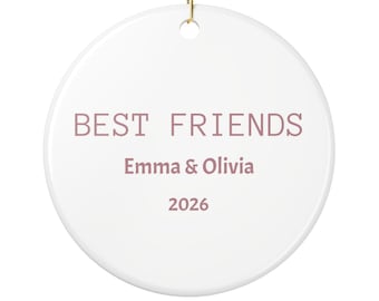 Op de afbeelding: Een rond wit keramieken ornament met de tekst BEST FRIENDS in een oudroze kleur, gevolgd door de namen Emma & Olivia en het jaartal 2026 in dezelfde kleur. Het ornament is voorzien van een gouden ophangkoord aan de bovenzijde.