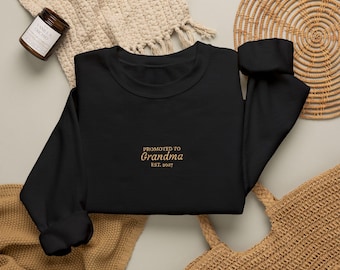 Op de afbeelding: Een zwart sweatshirt met lange mouwen en goudkleurig geborduurde tekst op de borst met de tekst PROMOTED TO Grandma EST. 2027. Het kledingstuk heeft een ronde hals en geribbelde boorden, plat neergelegd met neutrale accessoires.