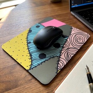 May include: A black computer mouse rests on a square mousepad featuring a patchwork design. The pattern consists of yellow, light blue, pink, and grey sections separated by black stitched lines, with polka dots and swirl motifs throughout.