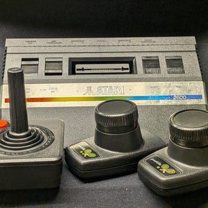 May include: A vintage Atari 2600 video game console with a joystick and two paddle controllers. The console is gray with the Atari logo and a rainbow stripe. The joystick has a red button.