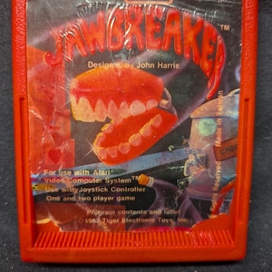 May include: A red plastic Atari video game cartridge for the game Jawbreaker by TigerVision. The label features a graphic of a set of teeth, the text "Jawbreaker," "Designed by John Harris," and "For use with Atari Video Computer System."