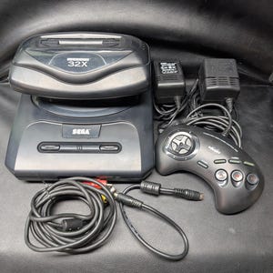 May include: A black Sega Genesis console with a 32X expansion unit attached, accompanied by a black wired game controller, two power adapters, and an AV cable with red, white, and yellow connectors. The console features the Sega logo.