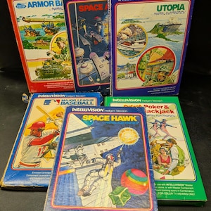 May include: A collection of vintage Intellivision video game cartridges. The boxes feature colorful graphics and titles like "Armor Battle," "Space Hawk," and "Utopia." The boxes are in various colors, including red, blue, and green.