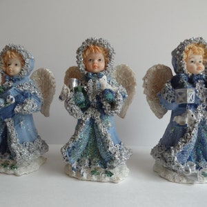 Pode incluir: Três figuras de anjos azuis e prateados com glitter e asas brancas. Cada anjo segura um item temático de Natal diferente.