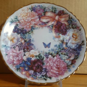 Puede incluir: Un plato de porcelana decorativo con un diseño de corona de flores. La corona está compuesta por flores rosas, moradas y blancas con hojas verdes. Una mariposa azul se encuentra en el centro de la corona.