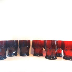 Peut inclure: Un ensemble de six verres à boire courts et cylindriques en verre rouge profond et translucide. Chaque verre présente un motif géométrique à facettes sur la moitié inférieure, créant un aspect texturé avec un bord lisse et clair.