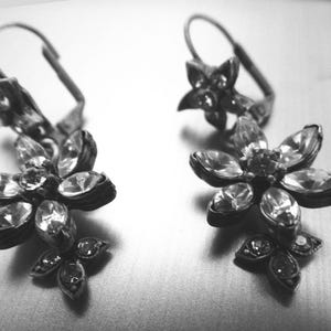 May include: A pair of silver-toned earrings with a floral design. Each earring features a flower shape with multiple clear, sparkling crystals. The earrings have a lever-back closure and are set against a neutral background.