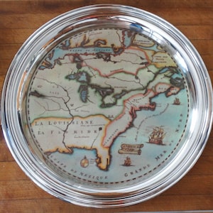 Könnte beinhalten: Ein runder Servierteller aus Silber mit einer detaillierten, farbenfrohen Karte von Nordamerika aus dem Jahr 1749. Die Karte enthält geografische Merkmale, Text und Illustrationen von Schiffen. Der Teller hat eine glänzende, reflektierende Oberfläche und einen breiten, erhöhten Rand.