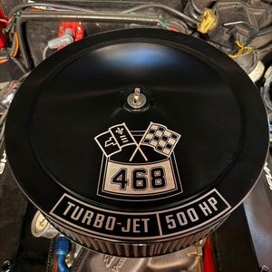 May include: A round black automotive air cleaner assembly featuring a central emblem with crossed checkered flags and the number 468. The base of the housing displays the white text TURBO-JET and 500 HP, positioned above a pleated filter element.