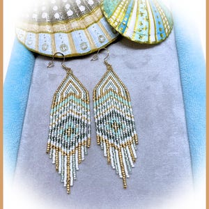 May include: A pair of long, dangling earrings crafted from small glass beads in a geometric pattern. The beads feature a color palette of white, mint green, metallic gold, and dark grey, arranged in a chevron design with a fringed bottom edge.