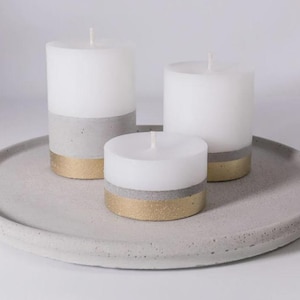 May include: Three white wax pillar candles with gray concrete bases and gold-painted bottom rims. The candles are arranged on a round, flat, gray concrete tray with a speckled texture and a raised edge.