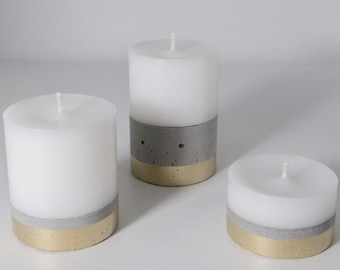 May include: Three cylindrical white wax candles of varying heights. Each candle features a base section composed of a gray concrete band topped with a metallic gold band. The candles have a single centered wick and a smooth wax finish.