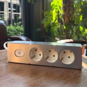 May include: A rectangular power strip with a brushed metal finish featuring three white electrical outlets and a circular power button with a power symbol. A white electrical cord extends from the left side of the unit.