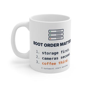 Op de afbeelding: Een witte keramische mok met een afbeelding van drie gestapelde serverpictogrammen en de tekst: BOOT ORDER MATTERS. 1. storage first. 2. cameras second. 3. coffee third. $ systemctl start morning. De tekst is in zwart en oranje.