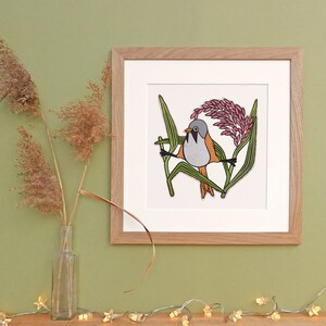 Könnte beinhalten: Ein gerahmter Druck mit einem stilisierten Vogel mit orangefarbenem, grauem und schwarzem Gefieder, der sich inmitten grüner Schilf und rosa Blüten befindet. Das Kunstwerk ist vor einem weißen Hintergrund in einem hellen Holzrahmen gesetzt. Getrocknete Gräser in einer Glasvase und sternförmige Lichterketten befinden sich im Vordergrund.